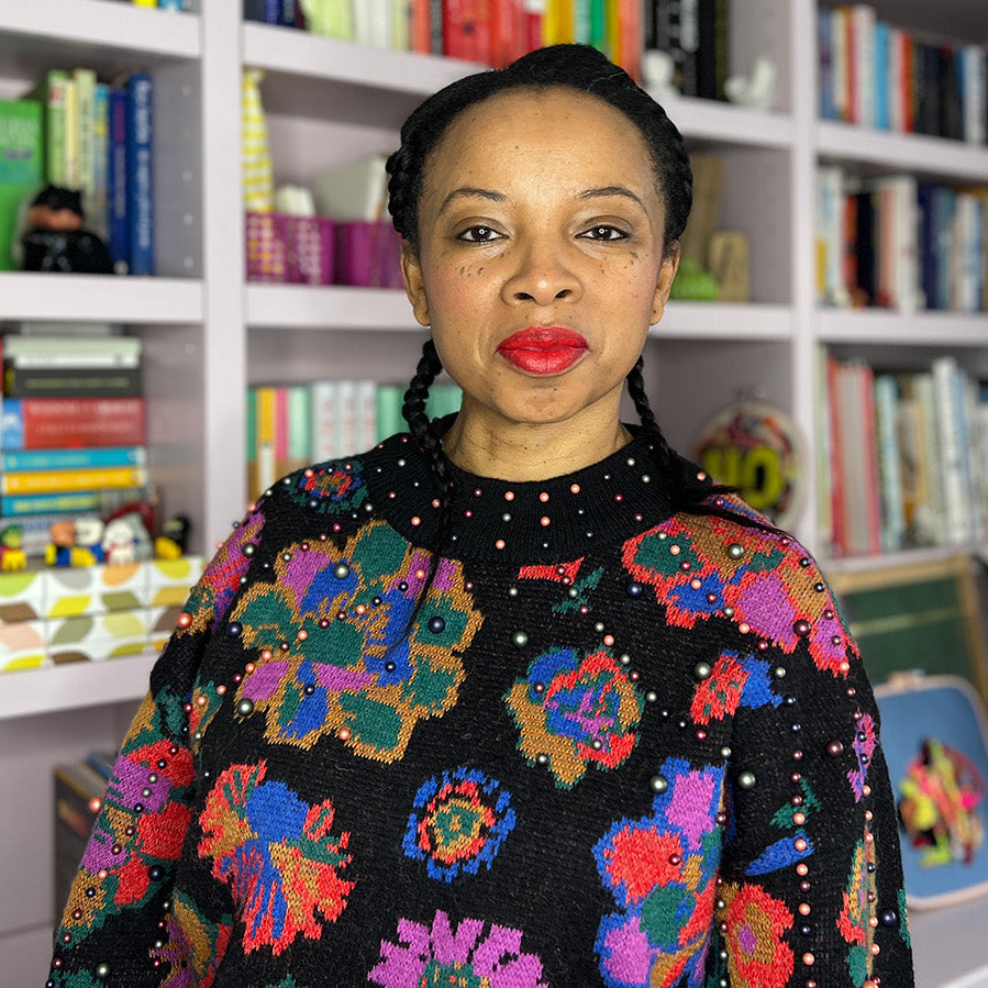 Natalie Lue with her hair in two braids wearing a bright, popping patterned Farm Rio jumper standing by her bookshelf in her studio